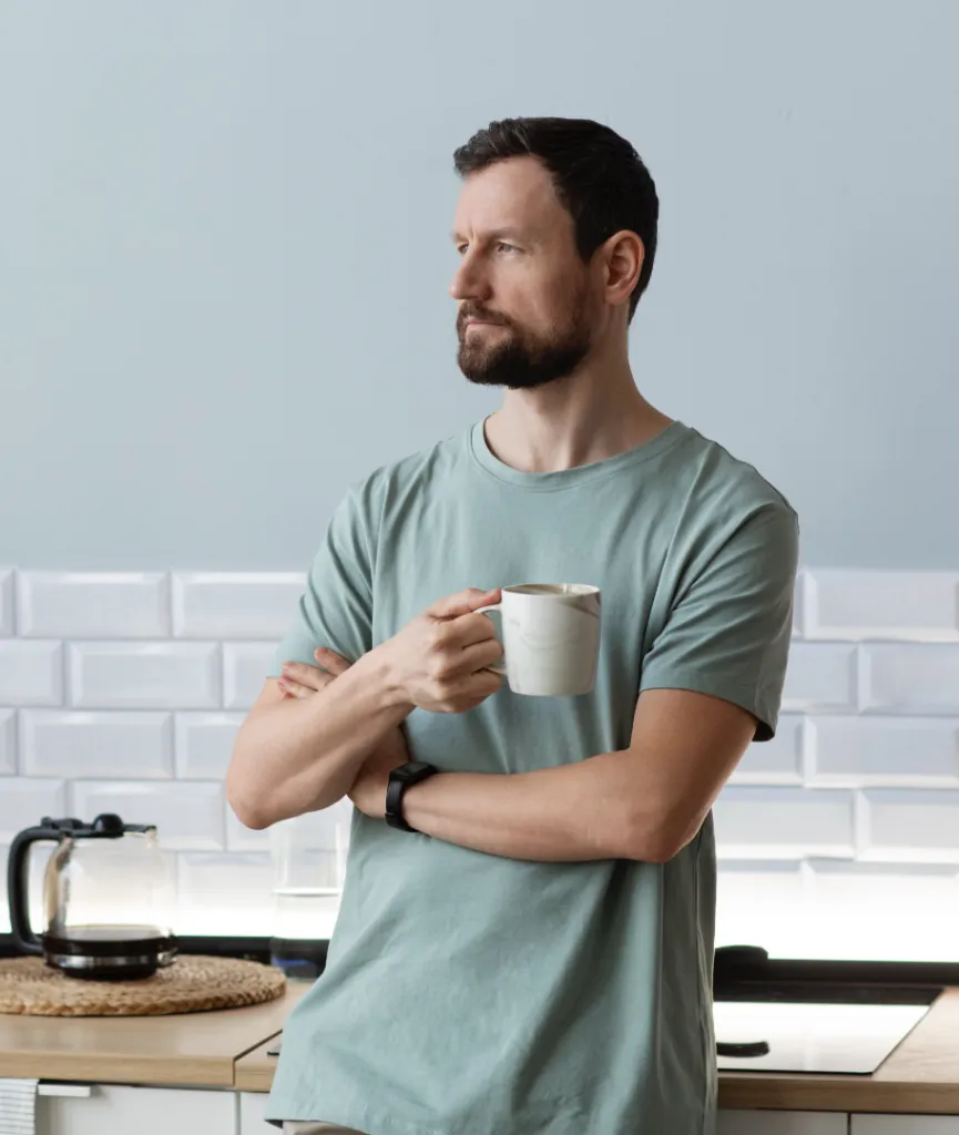 Hombre con barba en camiseta casual verde, sosteniendo una taza mientras parece reflexionar en una cocina moderna con una cafetera en el fondo.